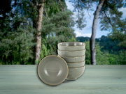 Stapel aus fünf gesprenkelten Keramikschalen in Beige auf einem hellen Holztisch vor unscharfem Waldhintergrund, eine Schale vorne liegend; Perspektive: leicht von vorne mit leicht erhöhter Ansicht.