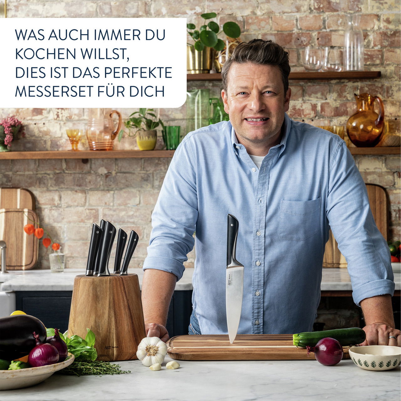 Mann in blauer Hemd steht in einer Küche vor einem Schneidebrett mit einem Messer darauf, umgeben von frischem Gemüse und einem Messerblock. Perspektive: Frontalansicht.