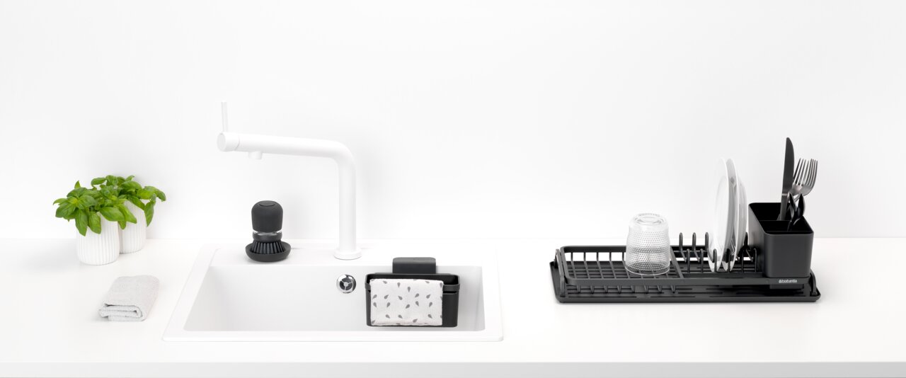 Moderne Küchenansicht mit Spül-Set, bestehend aus einem weißen Waschbecken mit schwarzem Spülmittelspender, einem schwarzen Schwammhalter und einem schwarzen Abtropfgestell mit Geschirr und Besteck, aus der Vogelperspektive.