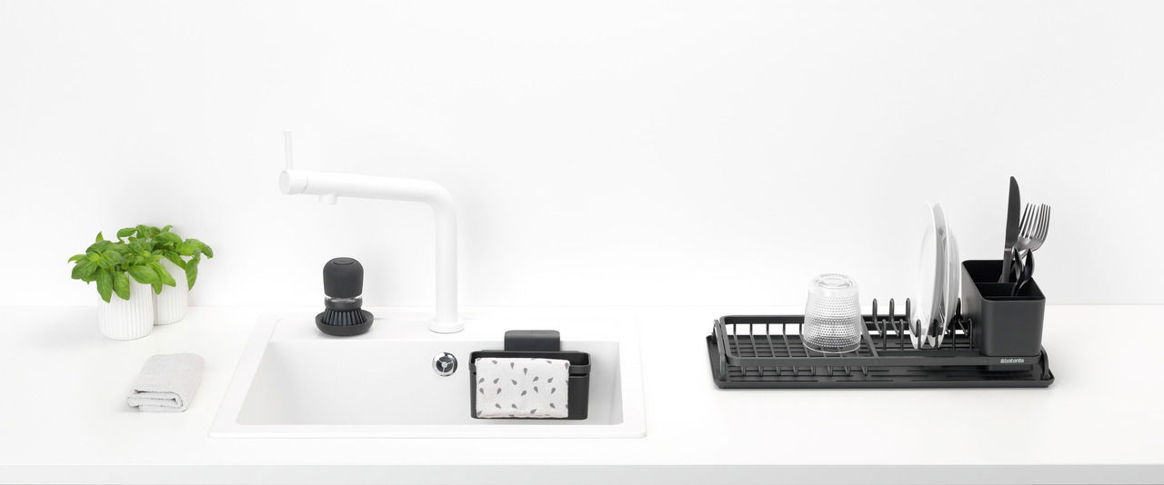 Brabantia Abtropfkorb-Set 4-tlg. SINKSIDE Moderne Küchenansicht mit Spül-Set, bestehend aus einem weißen Waschbecken mit schwarzem Spülmittelspender, einem schwarzen Schwammhalter und einem schwarzen Abtropfgestell mit Geschirr und Besteck, aus der Vogelperspektive.