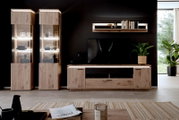Moderne entertainmentset voor de woonkamer van licht hout met twee verlichte vitrinekasten, een lowboard en een wandrek tegen een donkere muur, vooraanzicht