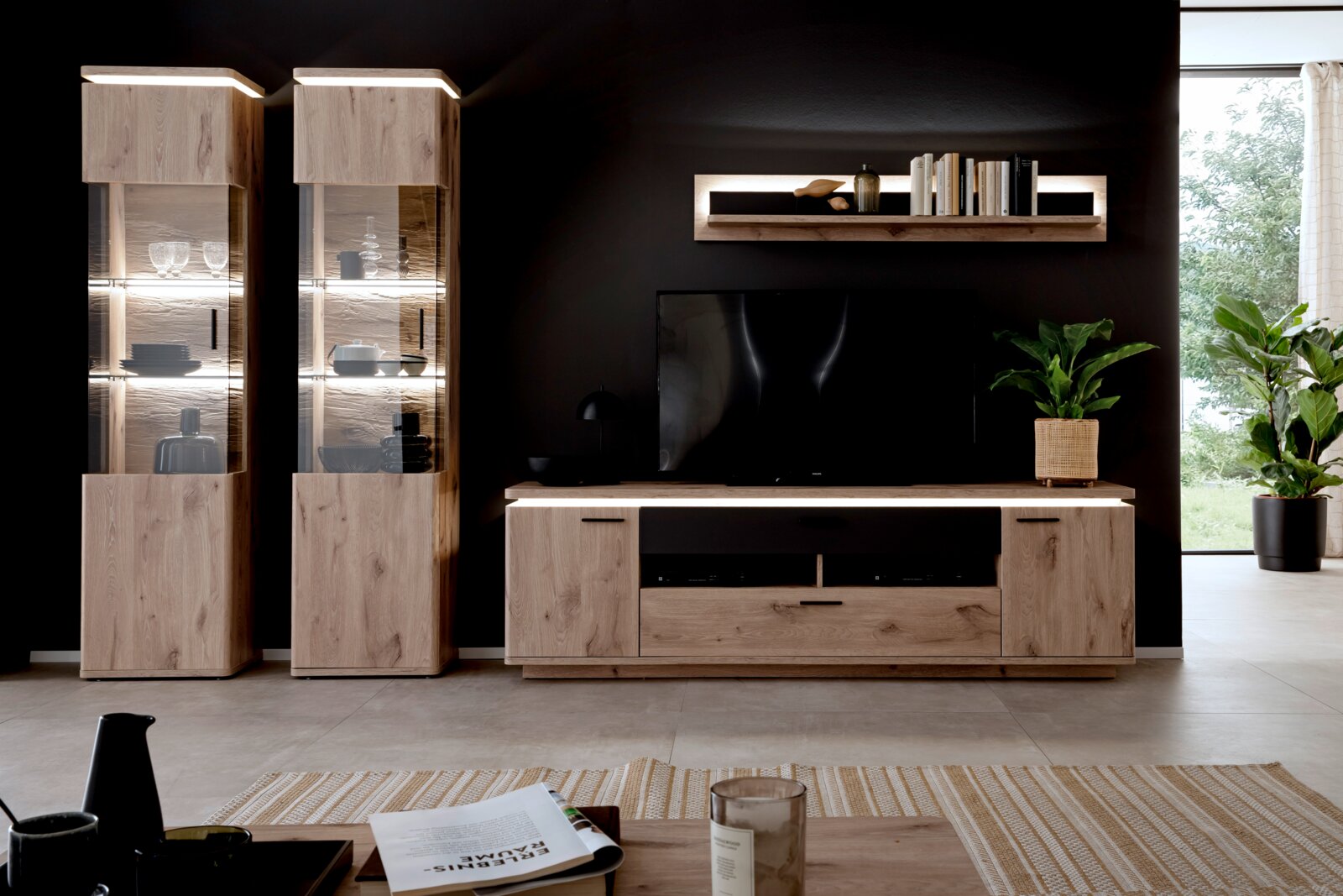 Moderne entertainmentset voor de woonkamer van licht hout met twee verlichte vitrinekasten, een lowboard en een wandrek tegen een donkere muur, vooraanzicht
