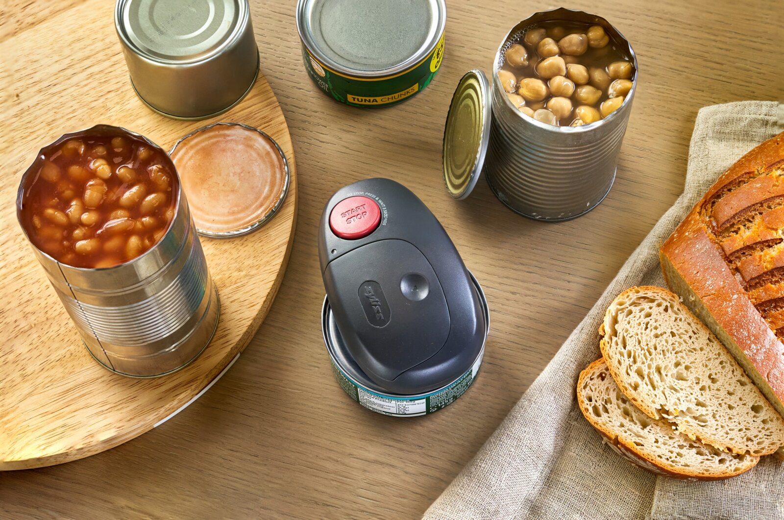 Elektrischer Dosenöffner in Schwarz mit roter Start/Stop-Taste auf einer Thunfischdose, umgeben von geöffneten Konservendosen mit Bohnen und Kichererbsen sowie Brot auf einem Holztisch, aufgenommen aus leicht schräger Vogelperspektive von oben.