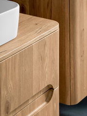 Detailaufnahme eines hellen Holz-Badmöbels mit abgerundeter Ecke und eingelassener Griffmulde, links oben ist der Rand eines weißen Aufsatzwaschbeckens zu sehen; Perspektive: Nahaufnahme in leichter Seitenansicht von oben.