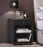 Schwarzer Nachttisch auf Rollen neben einem grauen Bett, mit Tischlampe, Bilderrahmen und modernem Wecker auf der Ablage sowie Büchern im offenen Fach, Ansicht schräg von vorn.