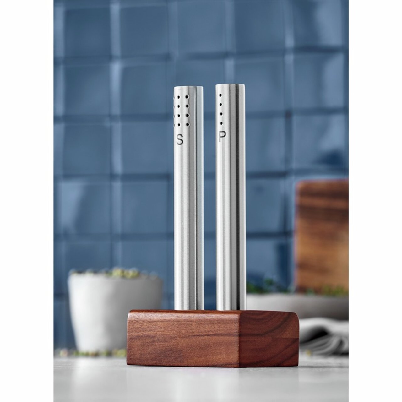 Modernes Salz- und Pfeffer-Set aus Edelstahl mit Holzständer, frontal abgebildet vor einer blauen Kachelwand.