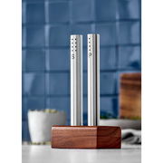 Modernes Salz- und Pfeffer-Set aus Edelstahl mit Holzständer, frontal abgebildet vor einer blauen Kachelwand.