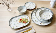 Een servies van keramiek met bloemmotief, schuin van bovenaf gefotografeerd: een ovaal serveerbord met salade, twee kommen en twee borden op een lichte houten tafel.