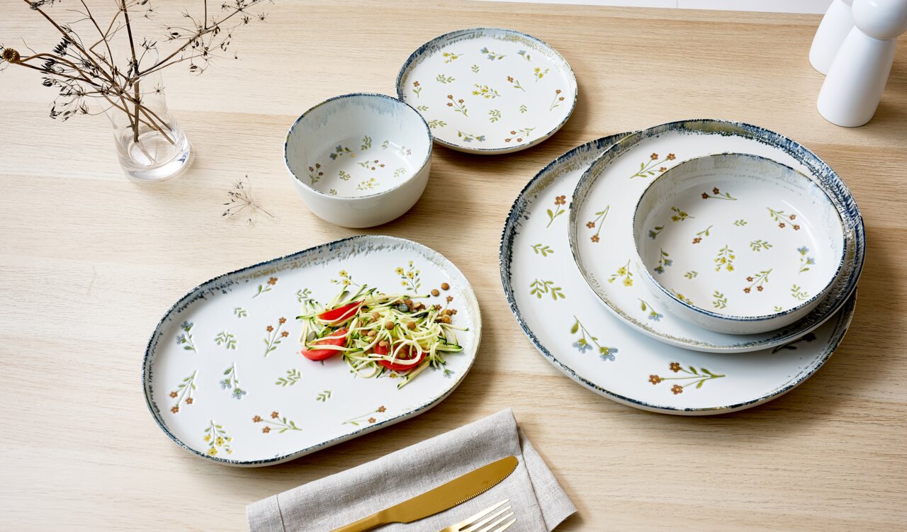 Een servies van keramiek met bloemmotief, schuin van bovenaf gefotografeerd: een ovaal serveerbord met salade, twee kommen en twee borden op een lichte houten tafel.