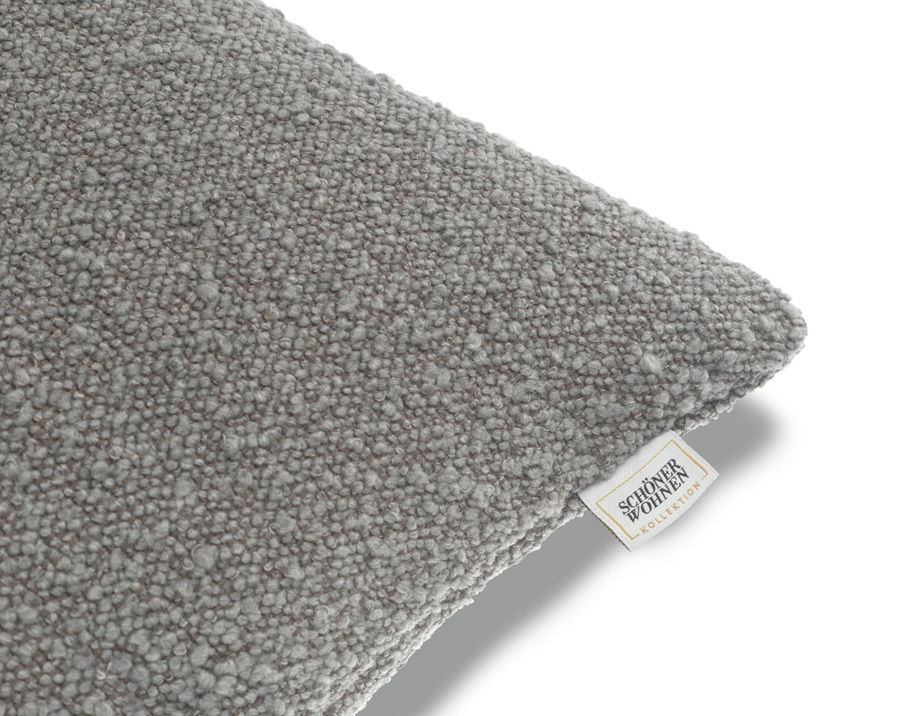 Graue Zierkissenhülle mit strukturierter Oberfläche, perspektivisch von der Seite fotografiert, mit sichtbarem Etikett der Schöner Wohnen Kollektion.