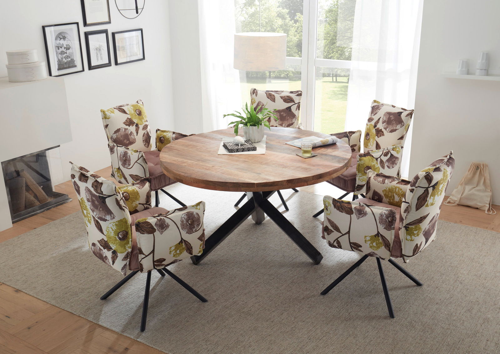 Runder Esstisch aus Holz mit schwarzem Metallgestell, umgeben von fünf gepolsterten Stühlen mit floralem Muster, in einem hellen Raum mit großen Fenstern, aus der Vogelperspektive betrachtet.
