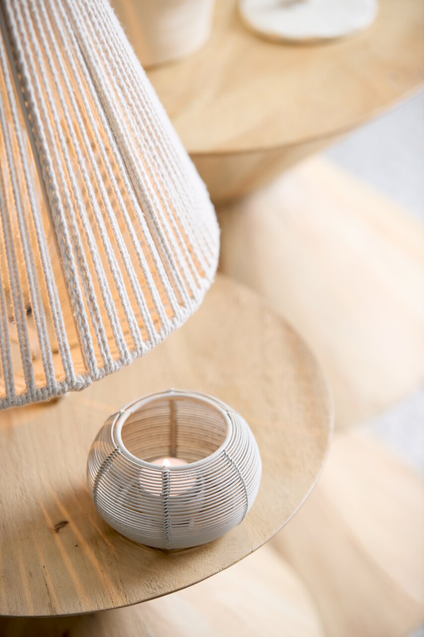 Theelichtje in een ronde, witte, matte houder van gevlochten materiaal, van bovenaf gefotografeerd op een houten tafel.