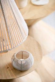 Theelichtje in een ronde, witte, matte houder van gevlochten materiaal, van bovenaf gefotografeerd op een houten tafel.