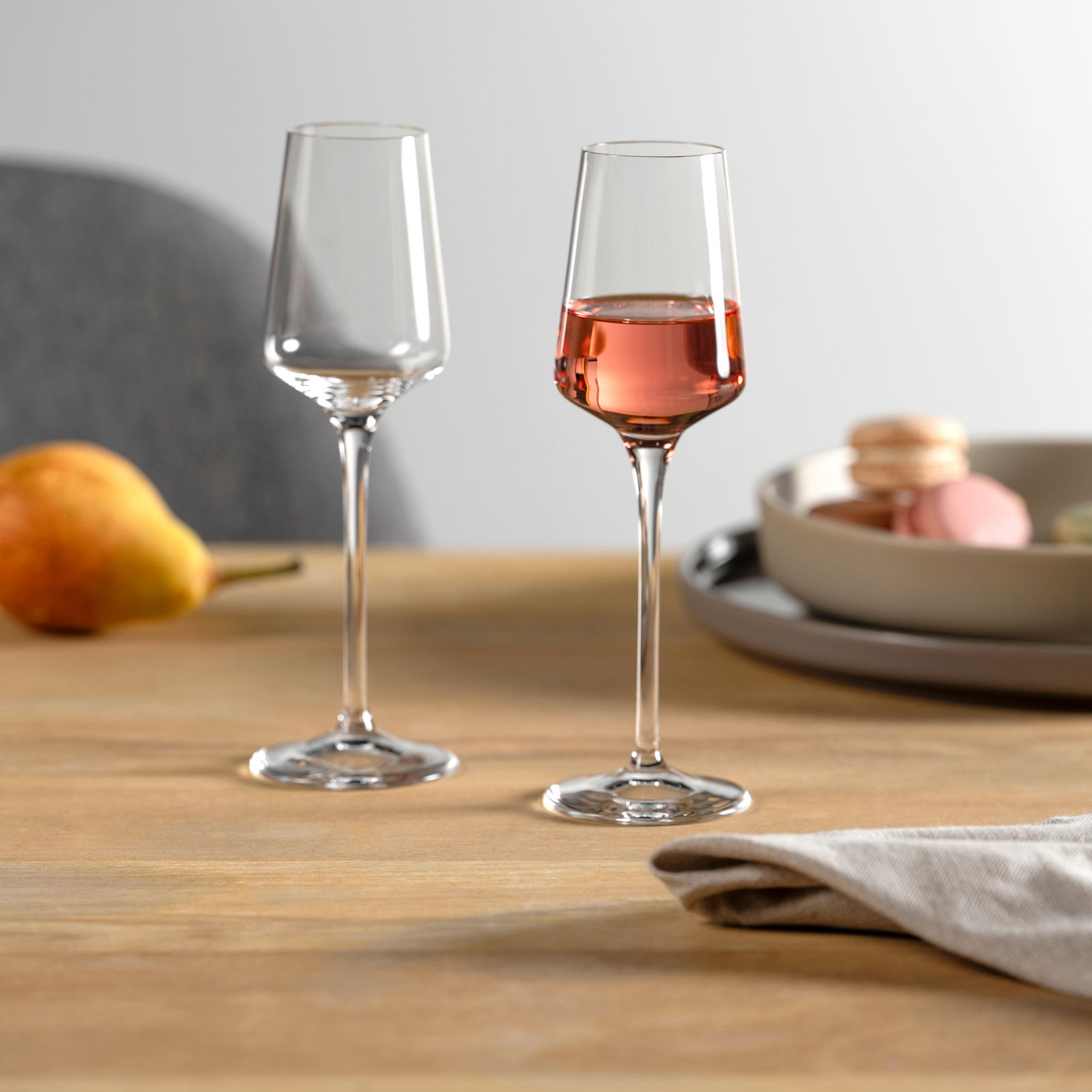 Zwei elegante Grappagläser auf einem Holztisch, eines leer und eines mit roséfarbenem Getränk gefüllt, im Hintergrund eine Birne und ein Teller mit Macarons, seitliche Perspektive.