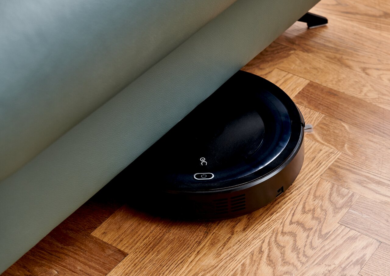 Ein Saugroboter, der unter einem Sofa auf einem Holzboden reinigt, aus seitlicher Perspektive.