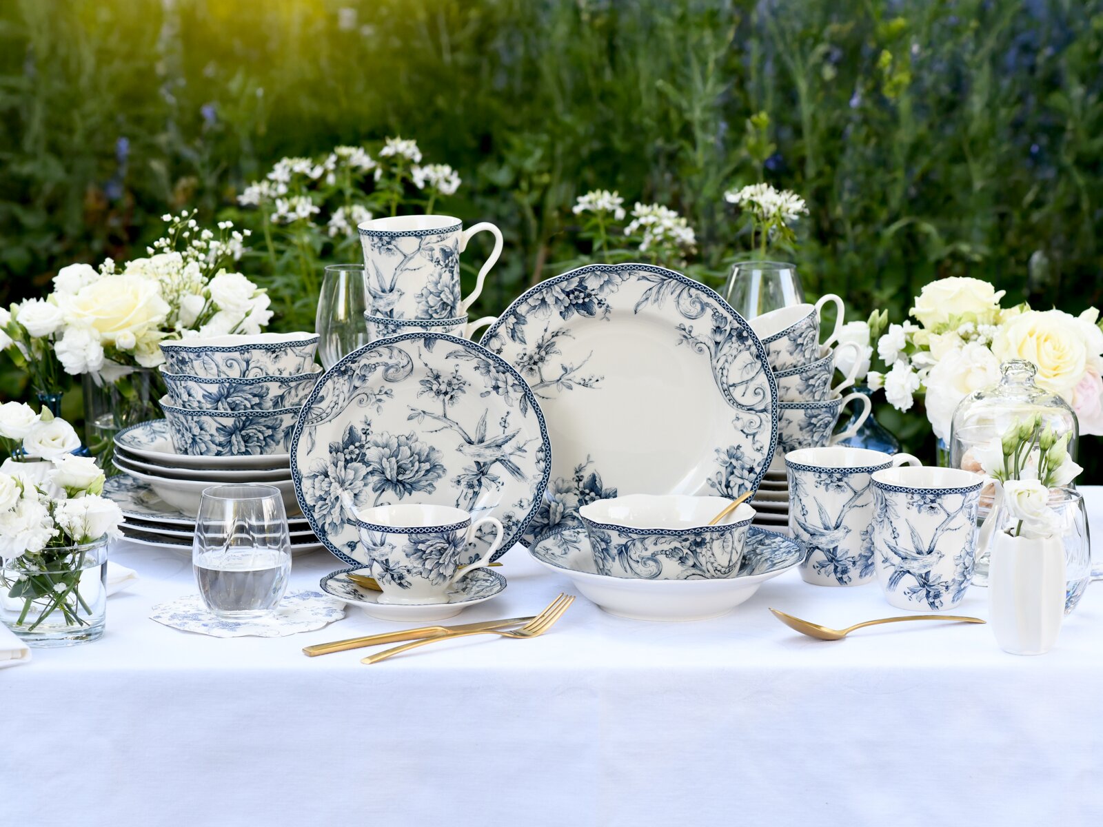Blau-weißes Porzellan-Geschirrset mit floralem Muster (Teller, Schalen, Tassen und Becher) dekorativ auf weißer Tischdecke im Garten arrangiert, fotografiert in frontaler Perspektive auf Augenhöhe.