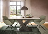 Moderner Esstisch mit X-förmigem Gestell und vier grünen Stühlen in einem hellen Raum mit großem Fenster, seitliche Perspektive.