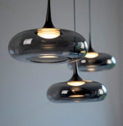 Moderne Pendelleuchte mit rauchgrauem, glänzendem Glas-Schirm in flacher Ovalform und warmem Licht, Nahaufnahme in leichter Untersicht/Frontansicht, weitere Lampen unscharf im Hintergrund