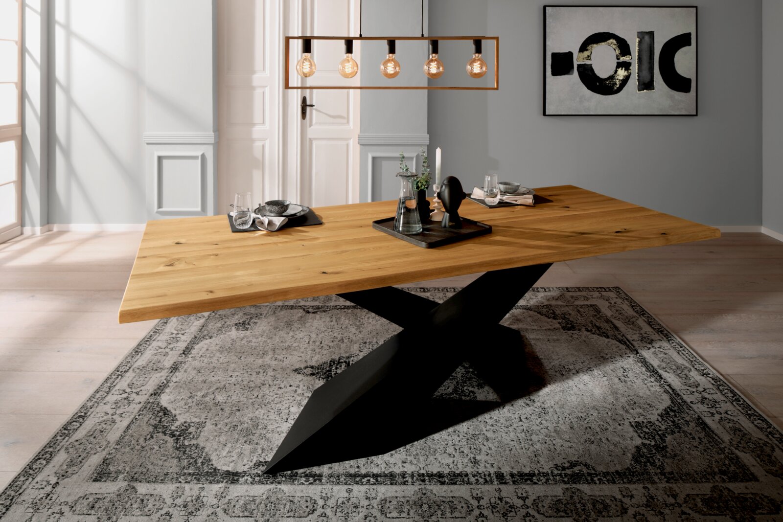 Moderner Esstisch aus Holz mit schwarzem Kreuzfußgestell, seitliche Perspektive, dekoriert mit Geschirr und Kerzen auf einem Teppich.
