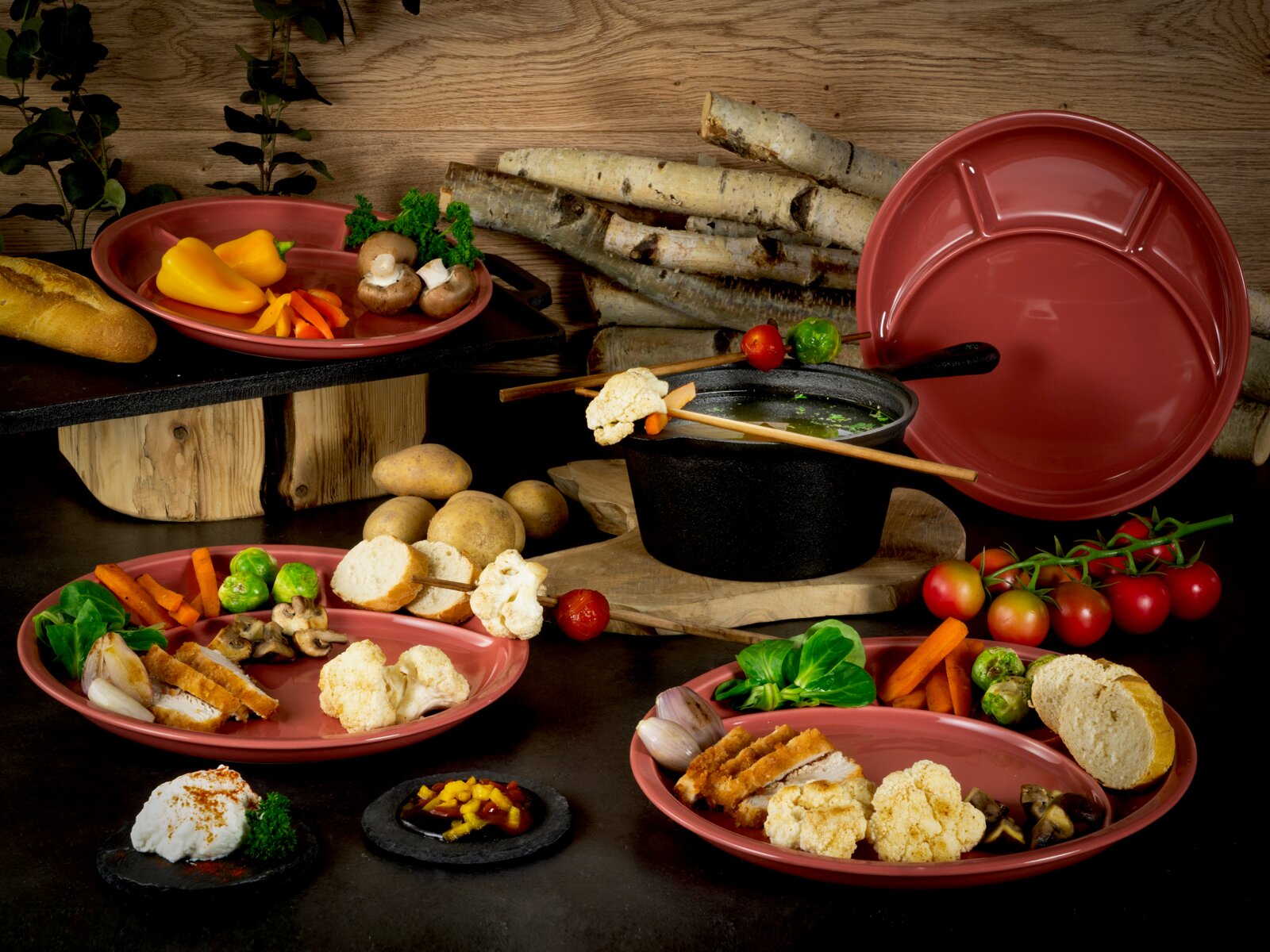 Rustikales Tisch-Arrangement mit rotem Keramik-Fondue-Set (Topf mit Griff, Teller und Platten) und Gemüse sowie Brot, fotografiert aus leicht erhöhter Frontperspektive.