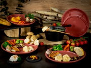 Rustikales Tisch-Arrangement mit rotem Keramik-Fondue-Set (Topf mit Griff, Teller und Platten) und Gemüse sowie Brot, fotografiert aus leicht erhöhter Frontperspektive.