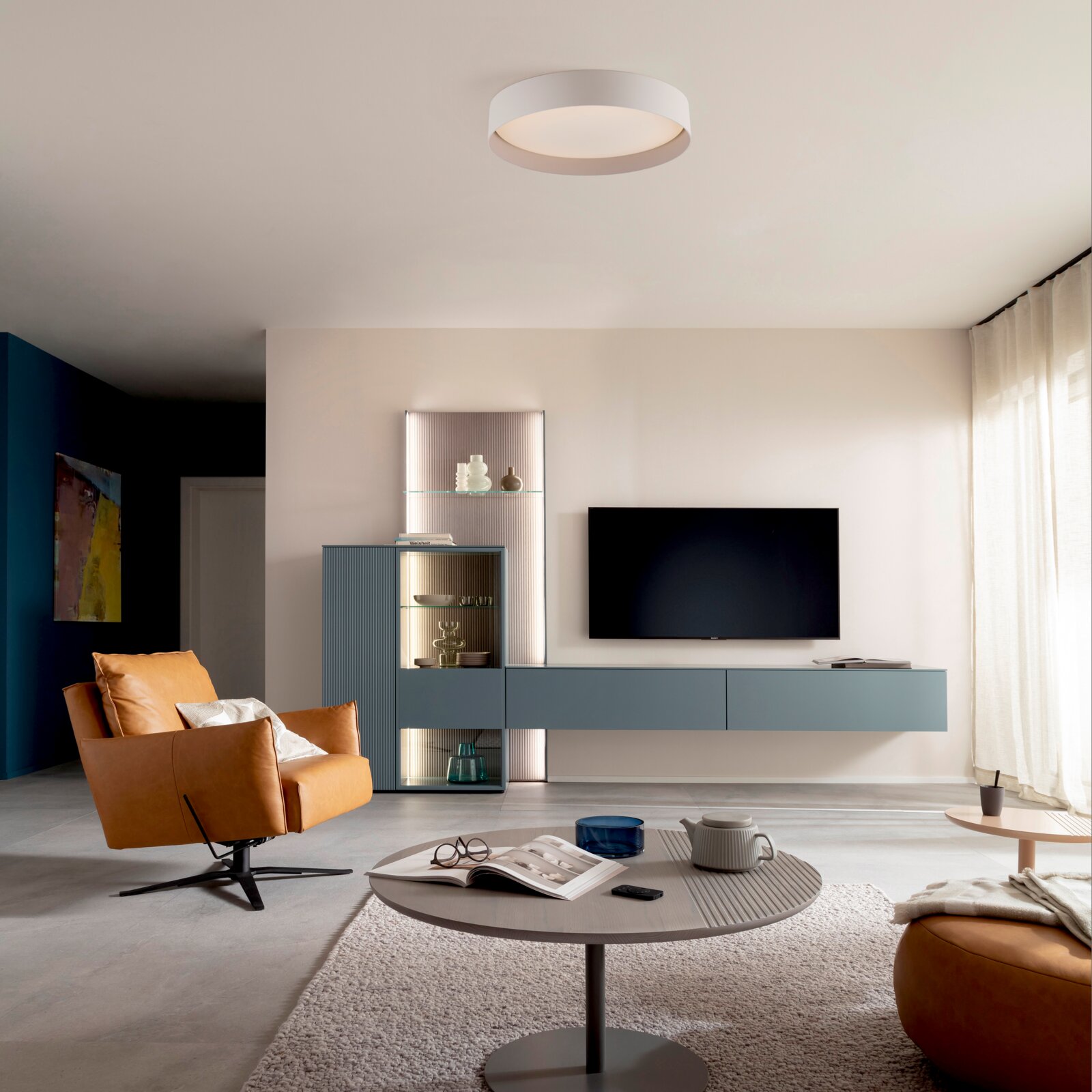Modernes Wohnzimmer mit weißer, runder LED-Deckenleuchte an der Decke, Blick aus der Raumecke nach oben/vorne darunter TV an der Wand, blaues Lowboard mit Vitrine, runder Couchtisch und cognacfarbener Sessel neben bodentiefen Vorhängen.