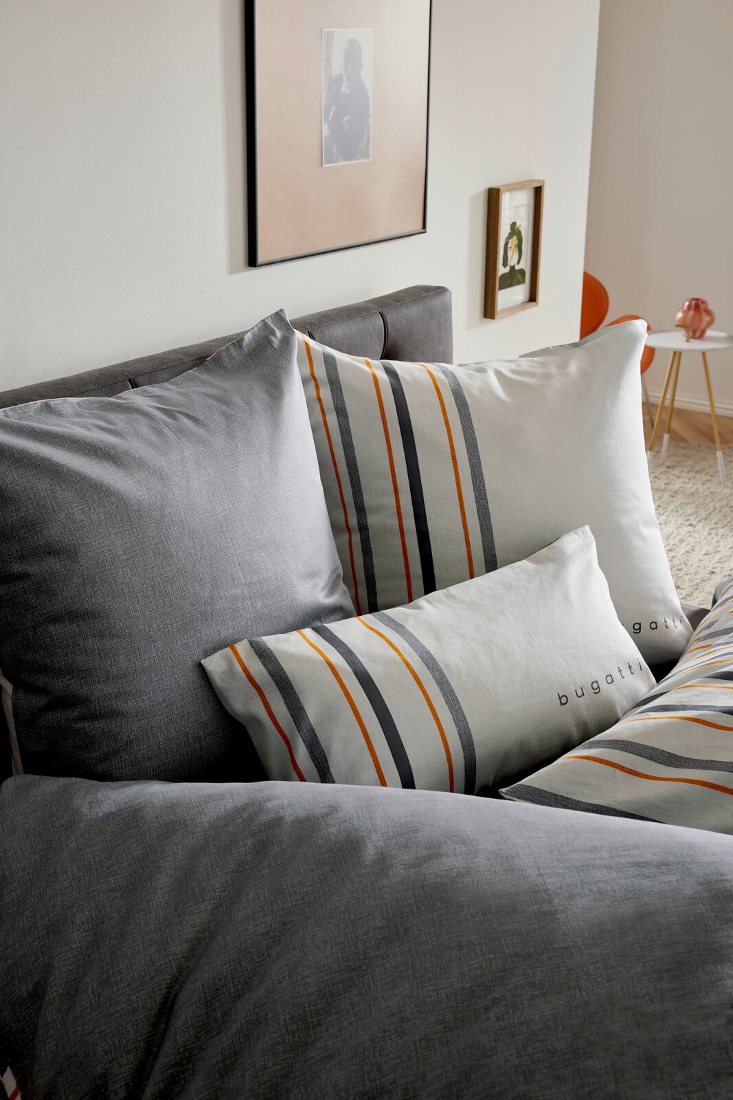 Schuin vooraanzicht van een grijs dekbedovertrek met bijpassende kussens, met wit en grijs gestreepte accenten met oranje lijnen en het bugatti-logo op de kussens, opgemaakt op een bed in een modern interieur.