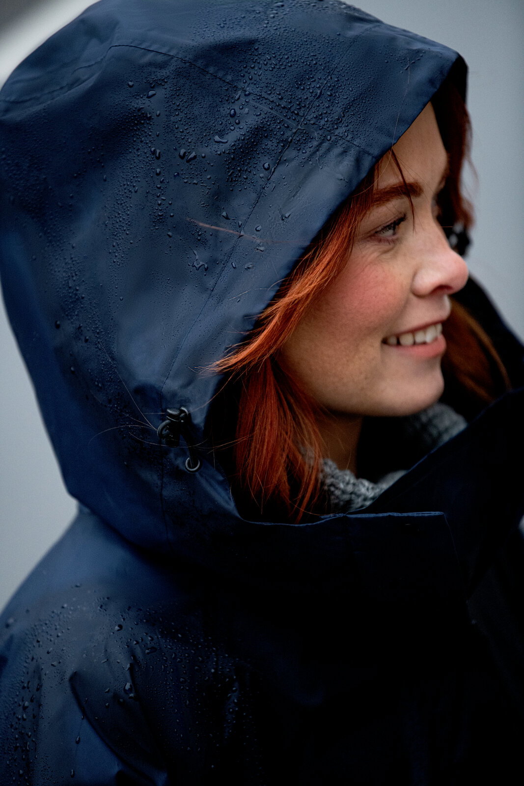 Nahaufnahme einer dunkelblauen, wasserabweisenden Regenjacke mit Kapuze und sichtbaren Regentropfen, getragen von einer Frau im Profil; Perspektive: seitliche Detailansicht (Close-up) der Kapuze.