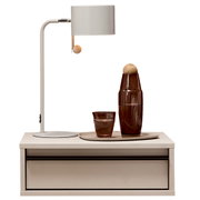 Frontansicht: moderne weiße Tischlampe mit zylindrischem Schirm und Holz-Zugschalter auf einem weißen Nachttisch; daneben braune Glasflasche und Glas auf Tablett