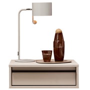 Frontansicht: moderne weiße Tischlampe mit zylindrischem Schirm und Holz-Zugschalter auf einem weißen Nachttisch; daneben braune Glasflasche und Glas auf Tablett