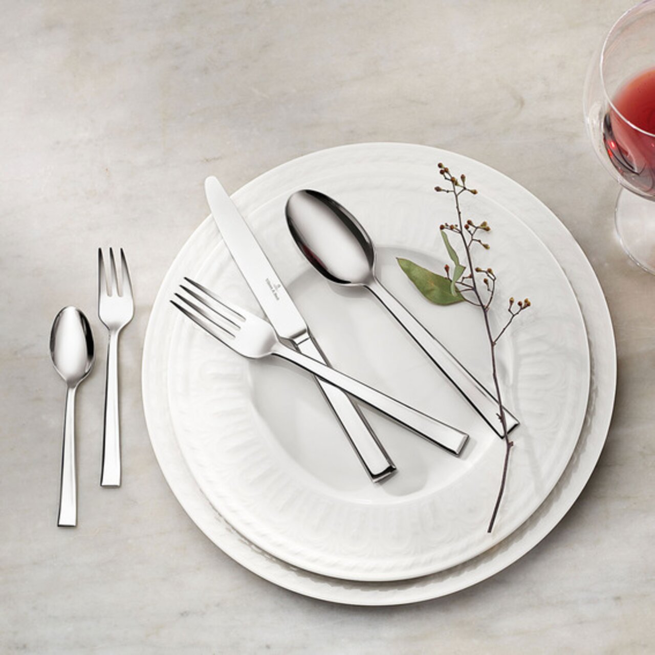 Elegantes 30-teiliges Tafelbesteck-Set Victor aus Edelstahl, auf einem weißen Teller arrangiert, mit einem Zweig und einem Glas Rotwein, aus der Vogelperspektive fotografiert.