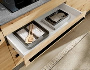Geöffnete Küchenschublade aus hellem Holz mit weißen Einsätzen, darin zwei Metalltabletts mit gefalteten Geschirrtüchern und Holz-Kochlöffeln; Perspektive: schräg von oben (Top-Down)