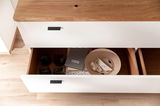 Offene Schublade eines TV-Lowboards aus Holz mit weißen Fronten, gefüllt mit Gläsern, einem gestreiften Notizbuch und Kabeln, aus der Vogelperspektive.