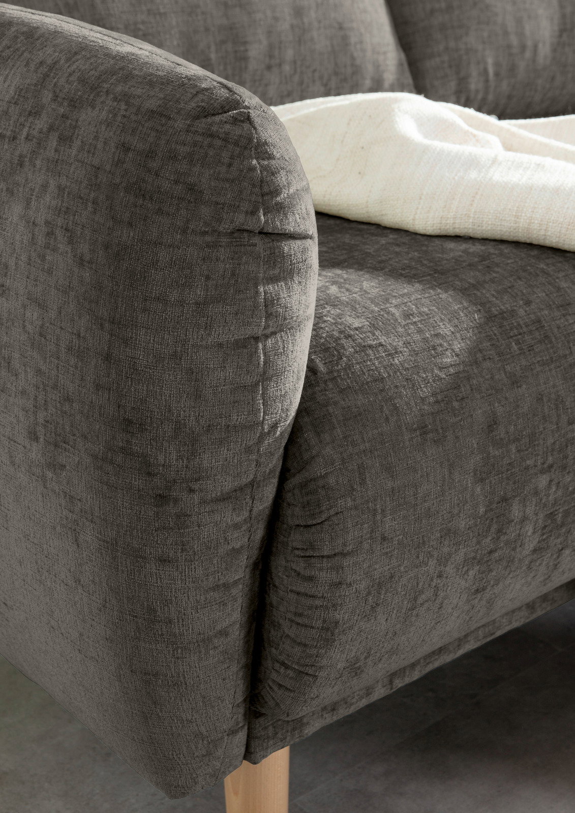 Nahaufnahme einer grauen Sofaecke mit Holzbeinen und einer weißen Decke, von der Seite betrachtet.