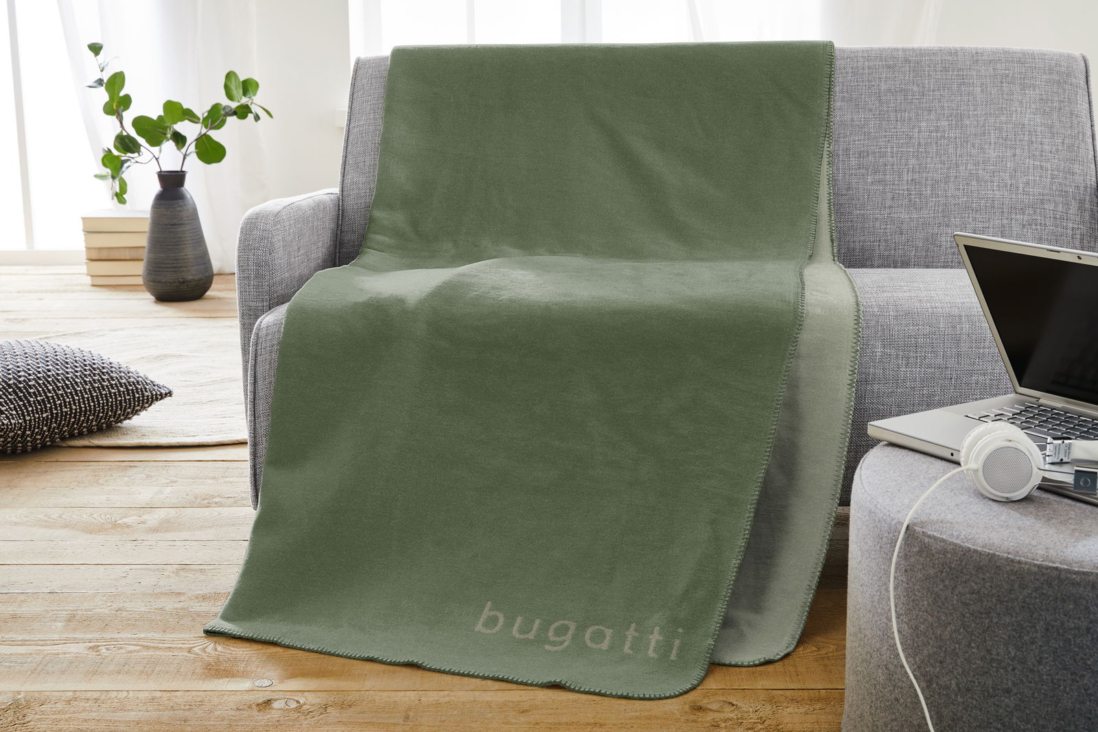 Grüne Jacquard Decke 150x200cm mit 'bugatti' Schriftzug, drapiert über einem grauen Sofa in einem modernen Wohnzimmer.