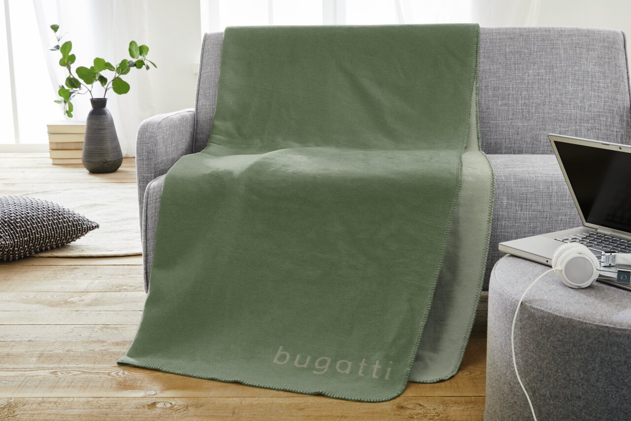 Bugatti Decke BUGATTI Grüne Jacquard Decke 150x200cm mit 'bugatti' Schriftzug, drapiert über einem grauen Sofa in einem modernen Wohnzimmer.