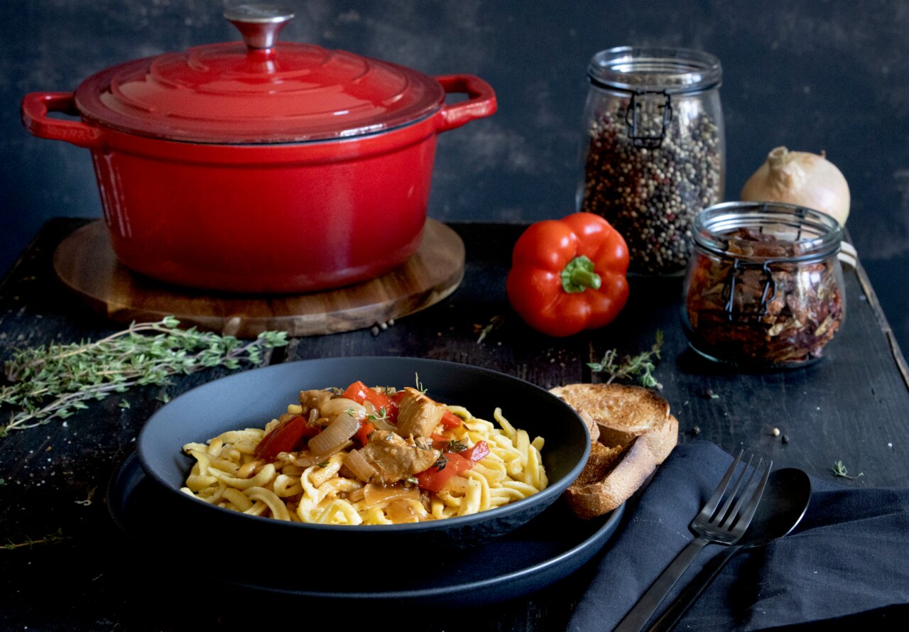 Roter runder Bräter aus Gusseisen, 26 cm, auf einem Tisch mit einer Schüssel Pasta im Vordergrund, fotografiert aus einer seitlichen Perspektive.