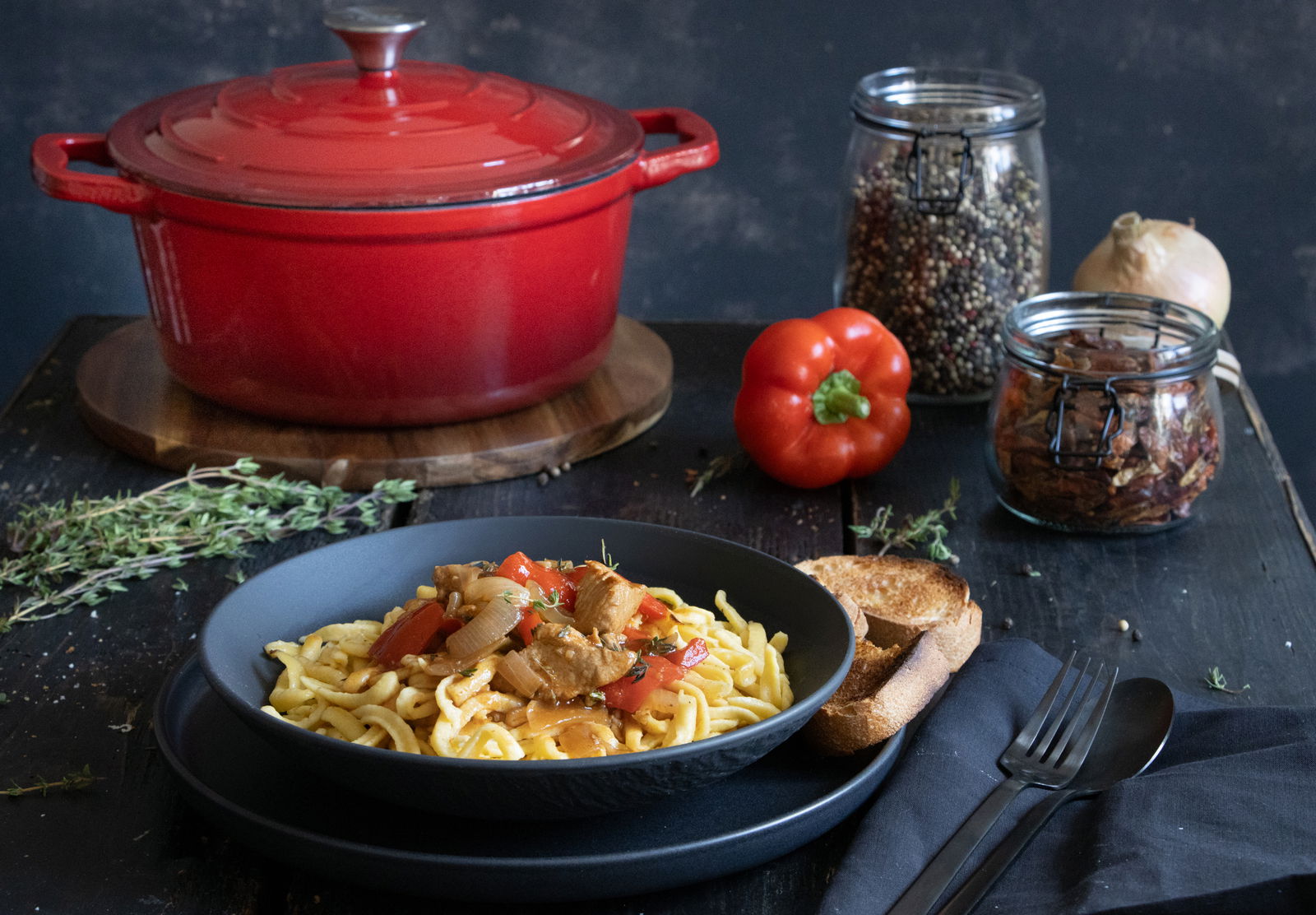 Roter runder Bräter aus Gusseisen, 26 cm, auf einem Tisch mit einer Schüssel Pasta im Vordergrund, fotografiert aus einer seitlichen Perspektive.