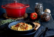 Rode ronde braadpan van gietijzer, 26 cm, op een tafel met een schaal pasta op de voorgrond, gefotografeerd vanuit een zijdelingse hoek.