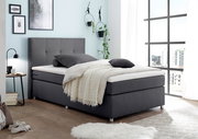 Modernes Boxspringbett in grauem Stoffbezug, seitliche Perspektive, dekoriert mit Kissen und Decke in einem hellen, stilvollen Schlafzimmer.