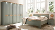 Helles Schlafzimmer-Set mit großem Bett, Nachttisch und mehrtürigem Kleiderschrank in Salbeigrün mit Holzdetails, fotografiert in schräger Frontansicht aus leicht erhöhter Perspektive.