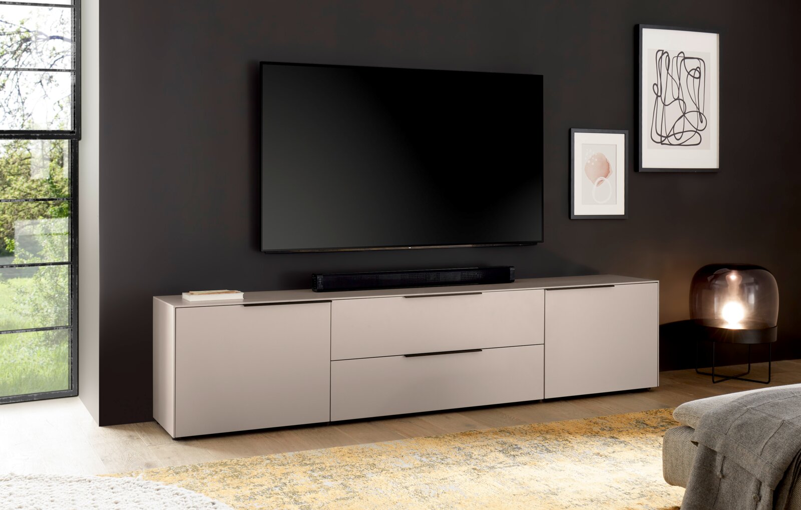 Modernes, niedriges TV-Lowboard/Sideboard in Hellgrau mit schwarzen Griffleisten unter einem wandmontierten Flachbild-TV im Wohnzimmer, frontale Perspektive mit leichter Seitenansicht.