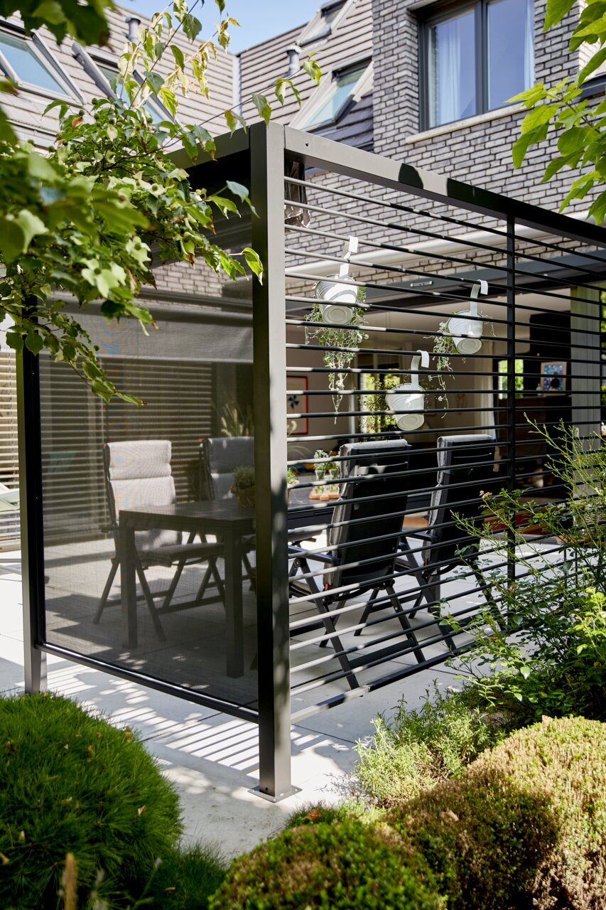 Moderne aluminium zichtscherm op een terras, zijaanzicht, omringd door planten en tuinmeubilair.