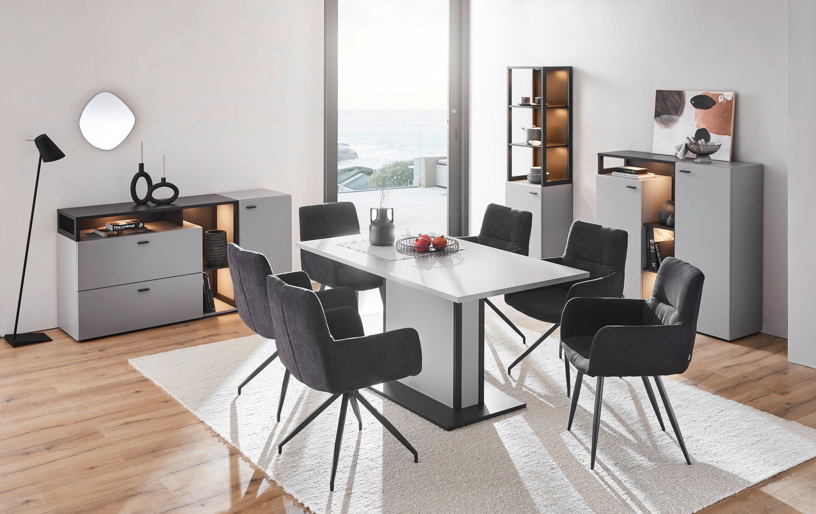 Moderne Esszimmermöbel in einem hellen Raum mit Meerblick, bestehend aus einem weißen Tisch, schwarzen Stühlen und grauen Sideboards. Perspektive: Frontalansicht.