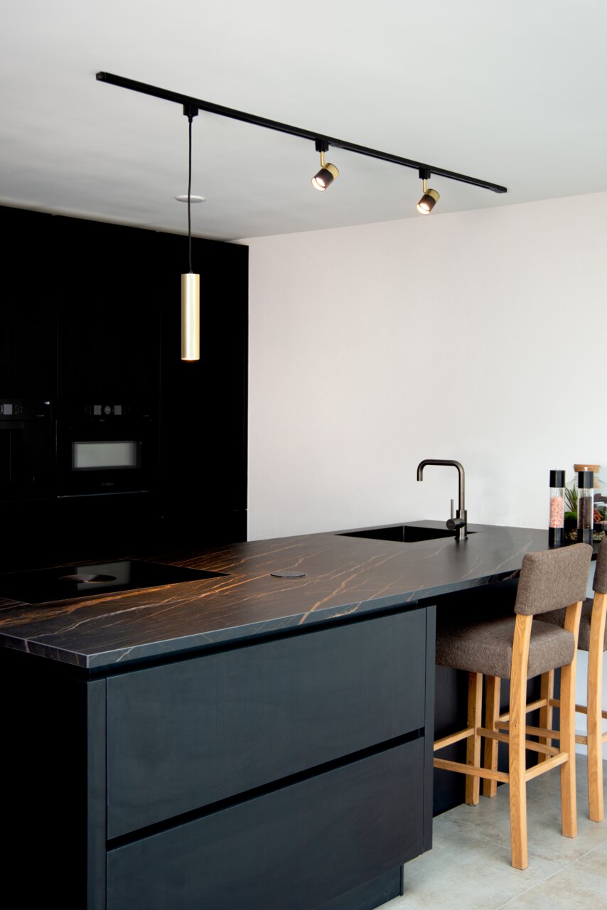 Moderne zwarte spotjes van het merk FLORIS, gemonteerd aan het plafond, verlichten een stijlvolle keuken met donkere kasten en een elegant werkblad. Aanzicht vanaf de zijkant.