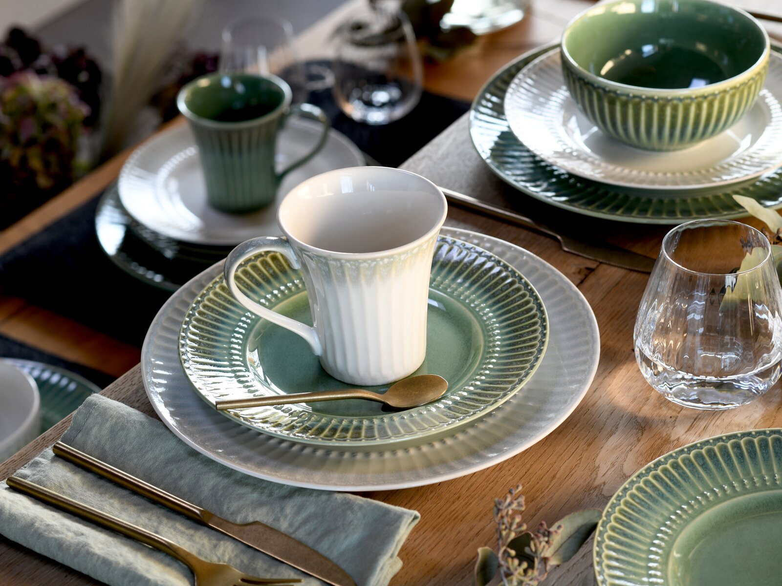 Gedeckter Holztisch mit grün-grauem Keramikgeschirr, weißer geriffelter Kaffeetasse auf grünem Unterteller, Schalen und Gläsern; Perspektive: leicht schräg von oben (3/4-Ansicht).
