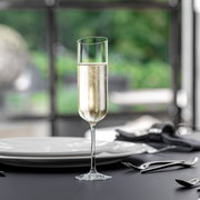 Op een gedekte tafel staat een elegante champagneglas, gevuld met bruisende champagne. Het perspectief is frontaal, met een onscherpe achtergrond die een rustige, groene omgeving toont.