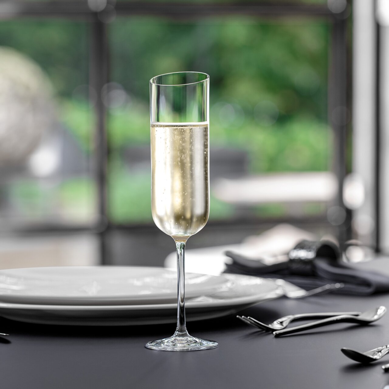 Op een gedekte tafel staat een elegante champagneglas, gevuld met bruisende champagne. Het perspectief is frontaal, met een onscherpe achtergrond die een rustige, groene omgeving toont.
