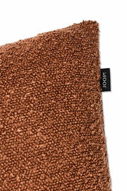 Nahaufnahme einer braunen Zierkissenhülle mit strukturierter Oberfläche und einem kleinen schwarzen Etikett an der Seite. Perspektive von der Ecke des Kissens.