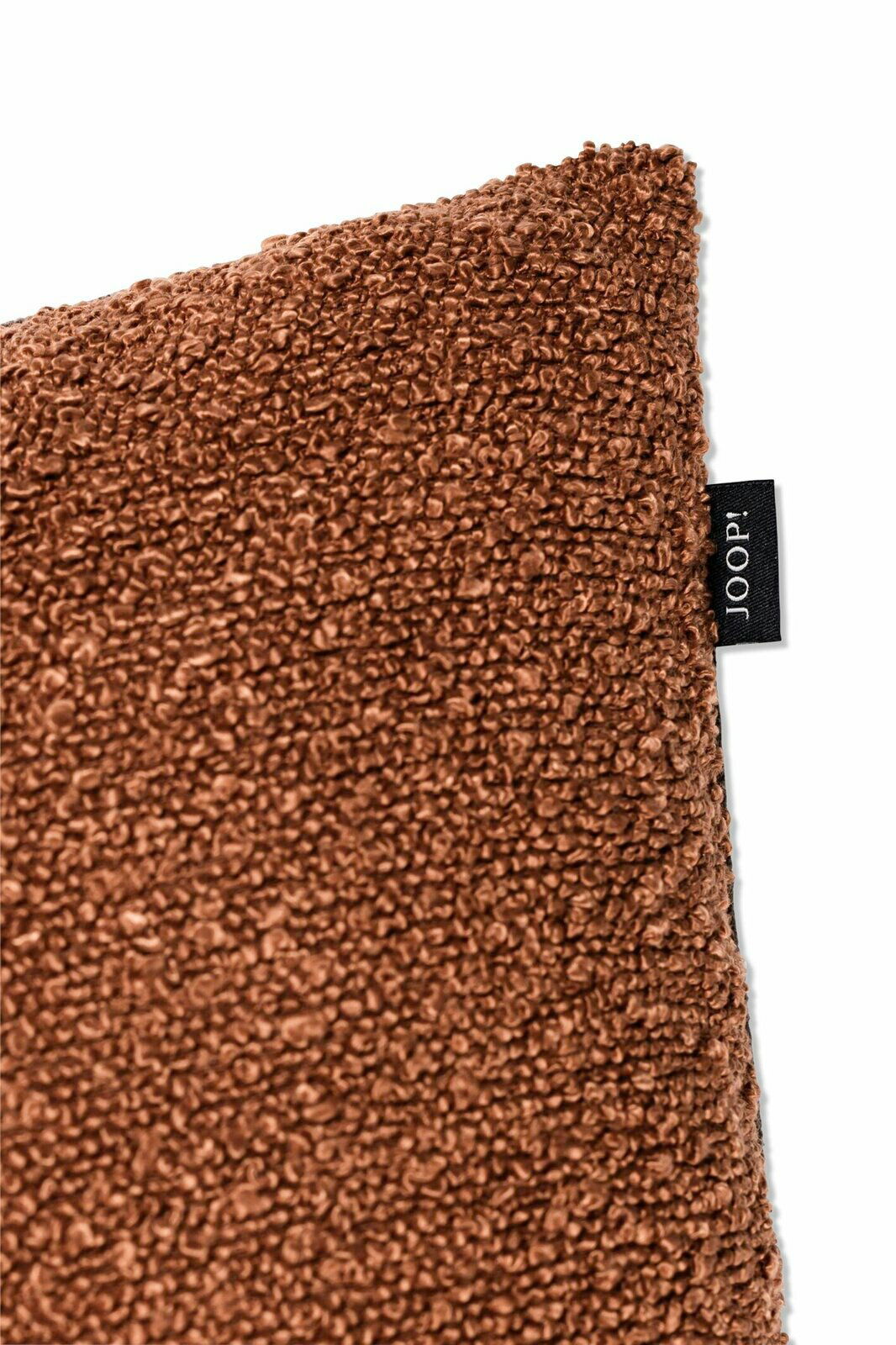 Nahaufnahme einer braunen Zierkissenhülle mit strukturierter Oberfläche und einem kleinen schwarzen Etikett an der Seite. Perspektive von der Ecke des Kissens.